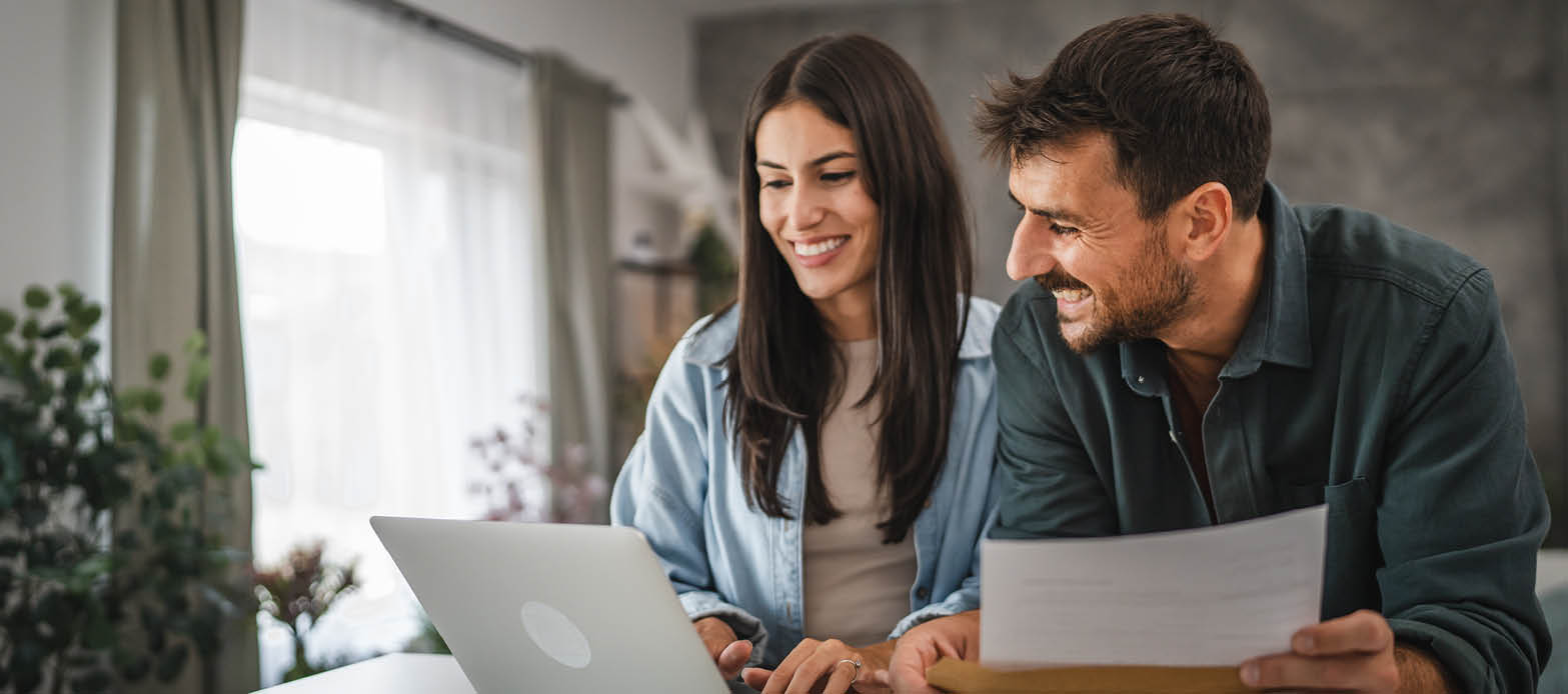A man and a woman smile and laugh as they look at a laptop and hold financial documents in their living room